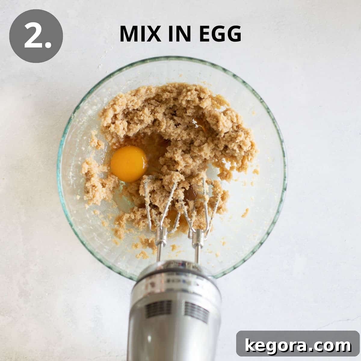 An egg being added to the creamed butter and sugar mixture in a glass mixing bowl, with a hand mixer poised above, ready to combine.