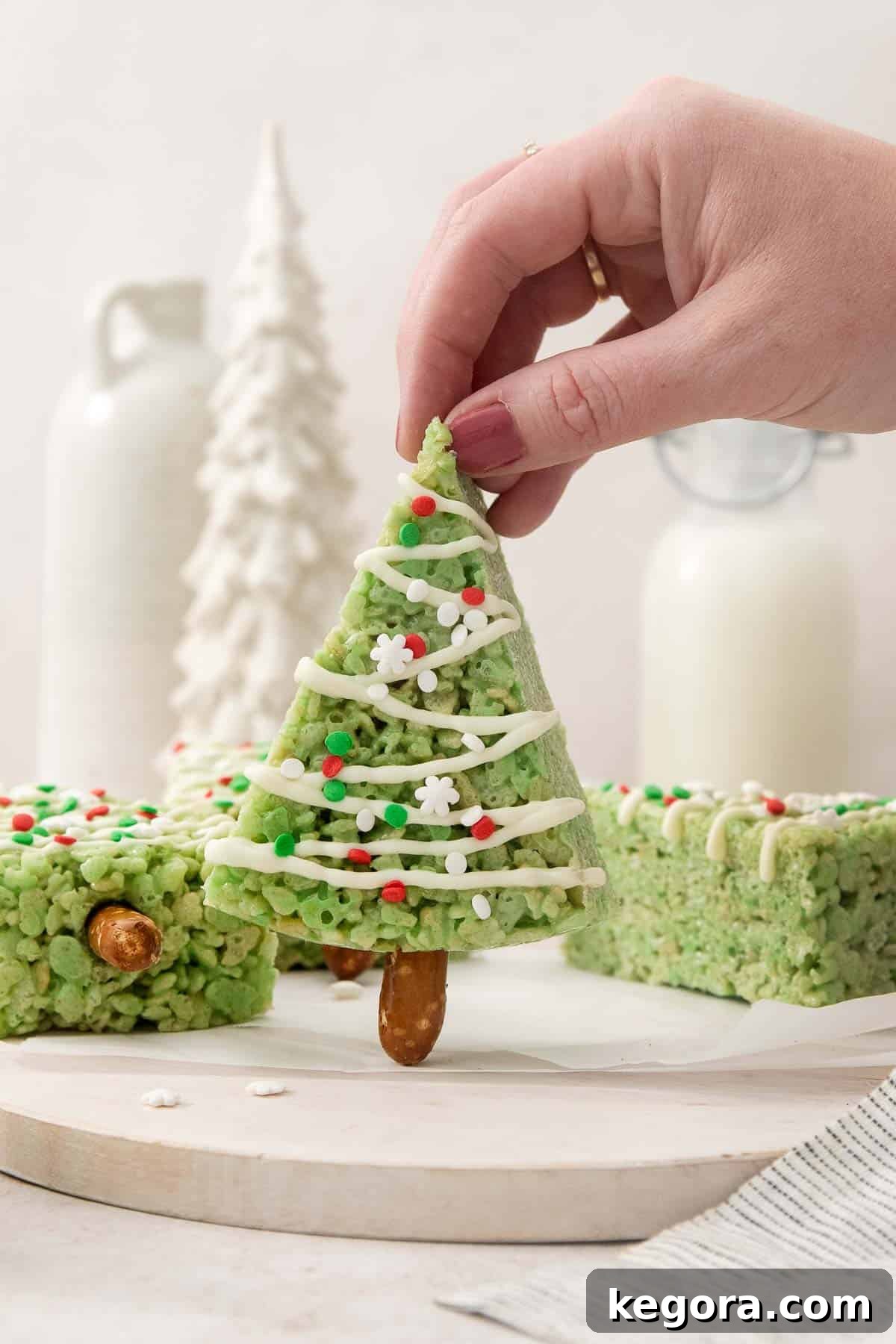 Sparkling Christmas Tree Bites 3 A hand holds one beautifully decorated Christmas tree crispy treat, with more festive treats blurred in the background on a snowy white platter.