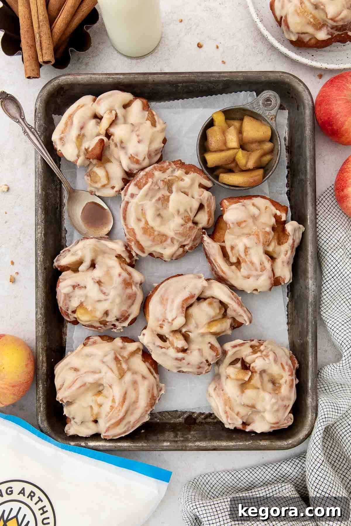 Crispy Gluten-Free Apple Delights 3 Beautifully glazed apple fritters arranged in a lined baking pan, surrounded by fresh apple slices and a spoonful of apple filling, highlighting their deliciousness.