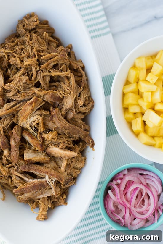 Overhead shot of shredded pineapple chipotle pork in a serving bowl, surrounded by fresh pineapple chunks and a smaller bowl of bright pink pickled onions, ready for taco assembly.