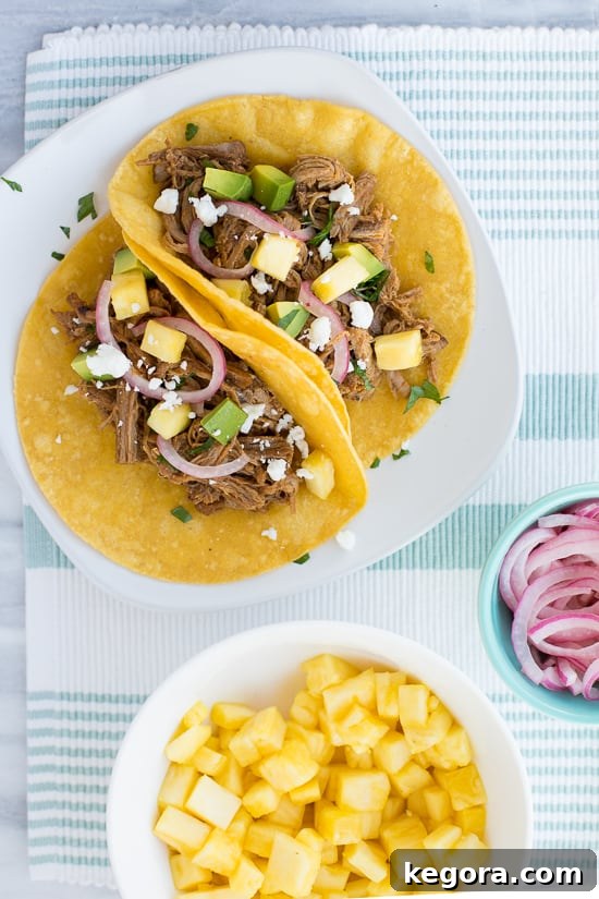 Overhead shot of three fully assembled pineapple chipotle pork tacos on a white plate, alongside a blue striped napkin. The tacos are topped with fresh pineapple and pickled red onions.