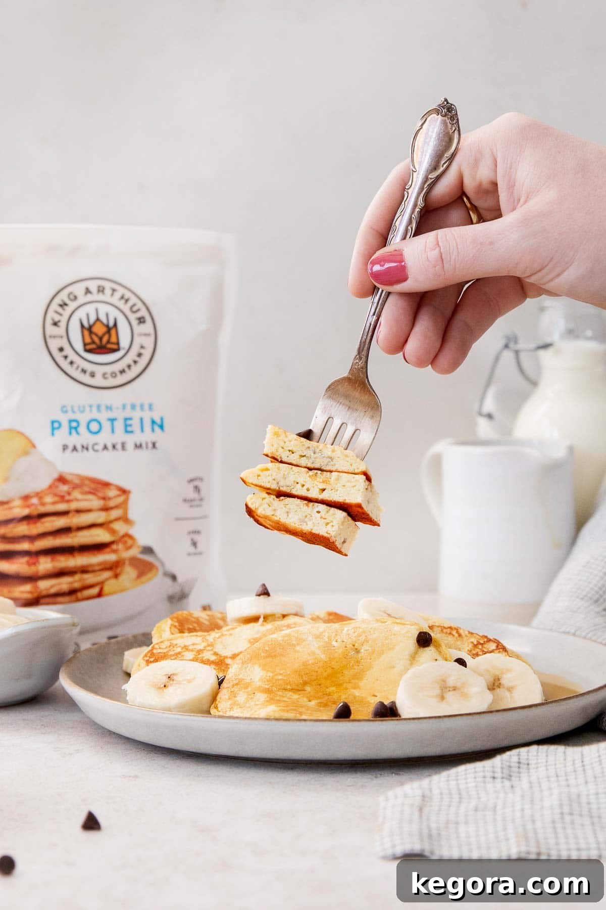 Protein Packed Gluten Free Dairy Free Banana Pancakes 6 Gluten-free banana protein pancakes on a plate, with a hand taking a bite with a fork and King Arthur Gluten-Free Protein Pancake mix in the background.