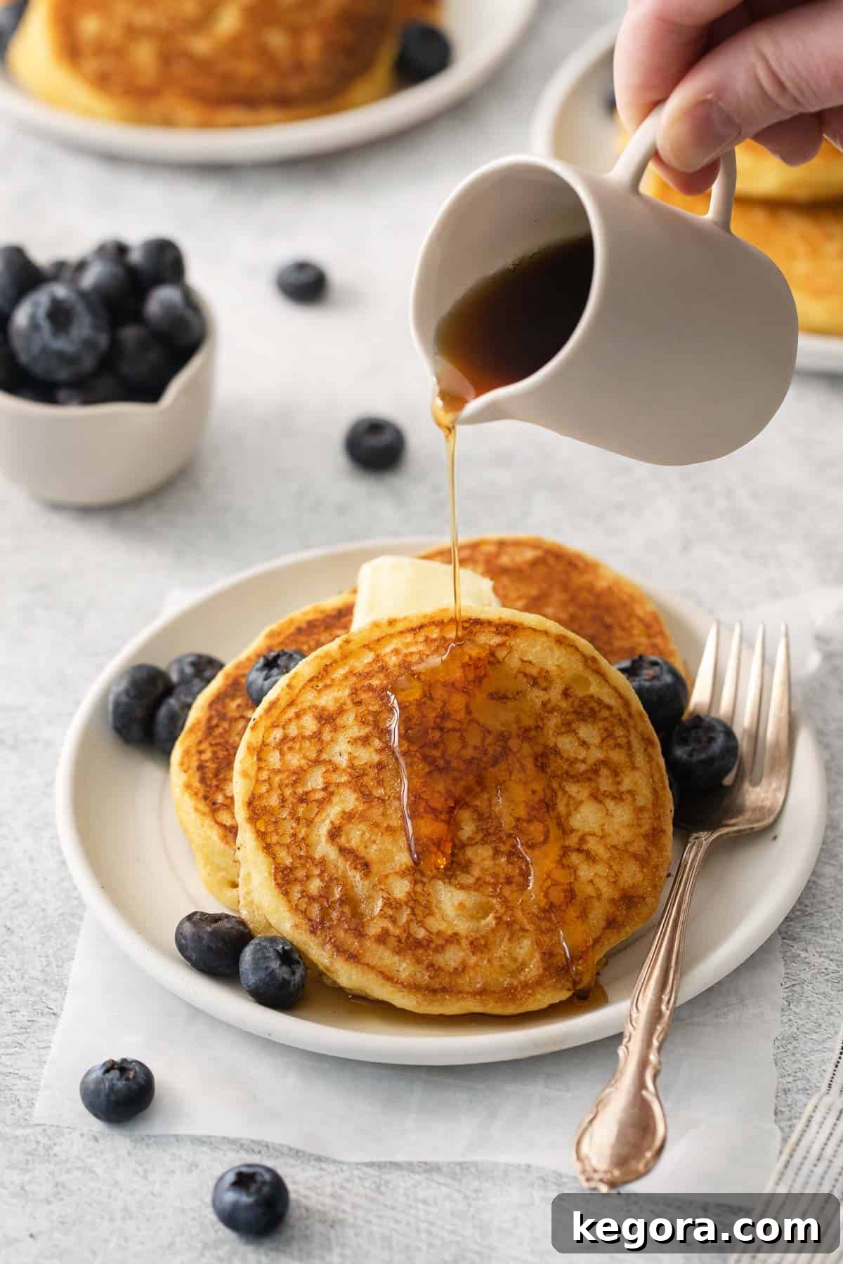 Silky Corn Cakes 2 Golden cornmeal pancakes stacked on a plate, with a hand pouring rich maple syrup over the top.