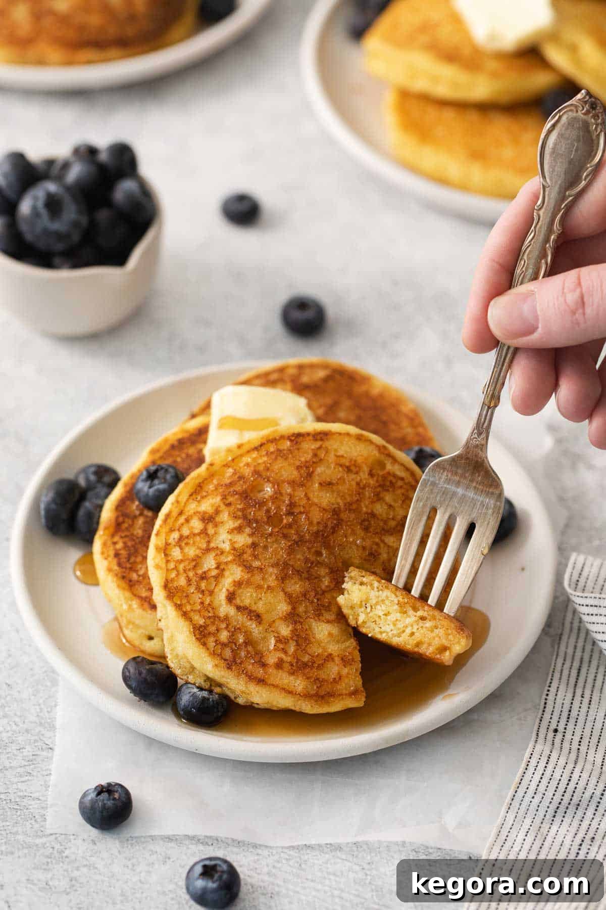 Silky Corn Cakes 10 A fluffy cornmeal pancake on a white plate with a fork taking a bite, showing the tender texture and golden interior.