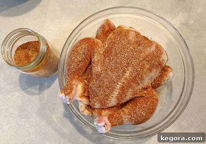 Raw whole chicken in a glass mixing bowl with spice rub covering it, ready for seasoning
