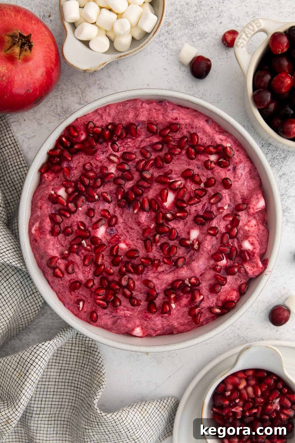 Vegan Cranberry Cloud 3 A large bowl of dairy-free cranberry fluff on a kitchen countertop, with a whole pomegranate in the soft-focus background, ready to be served at Thanksgiving or Christmas dinner.