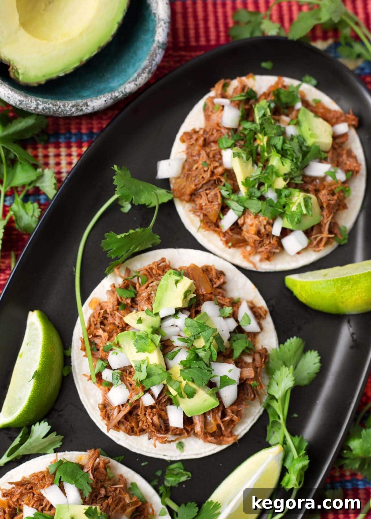 Barbacoa Beef, tender and garnished with fresh avocado, onion, and cilantro, beautifully arranged on white corn tortillas and served on a sleek black oblong platter.