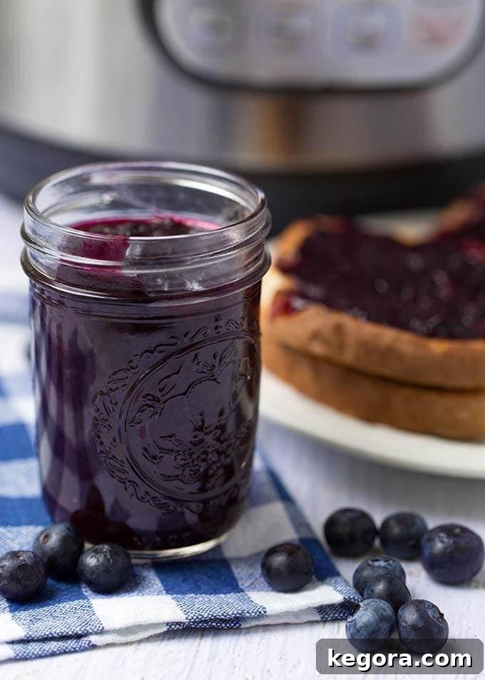 Homemade Instant Pot Blueberry Jam 5 A mason jar filled with vibrant blueberry jam sitting on a wooden surface, with a slice of bread spread with jam and an Instant Pot blurred in the background.