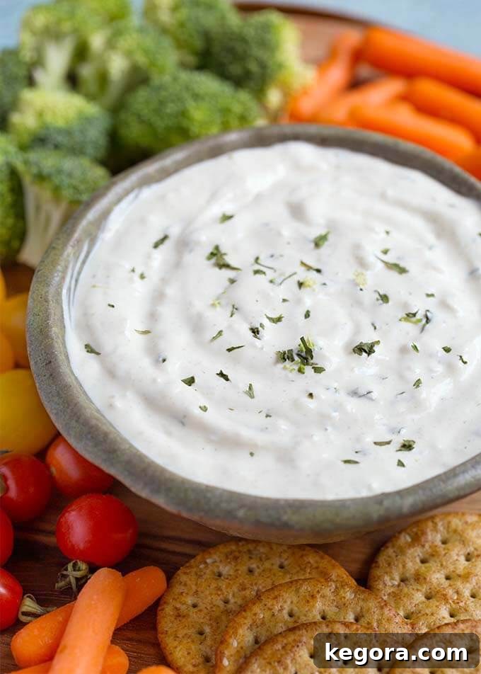Close-up view of the smooth, creamy texture of Ranch Vegetable Dip in a gray bowl, garnished with a sprinkle of fresh herbs. It sits on a round wooden board, surrounded by crisp, colorful vegetables and gourmet crackers, inviting indulgence.