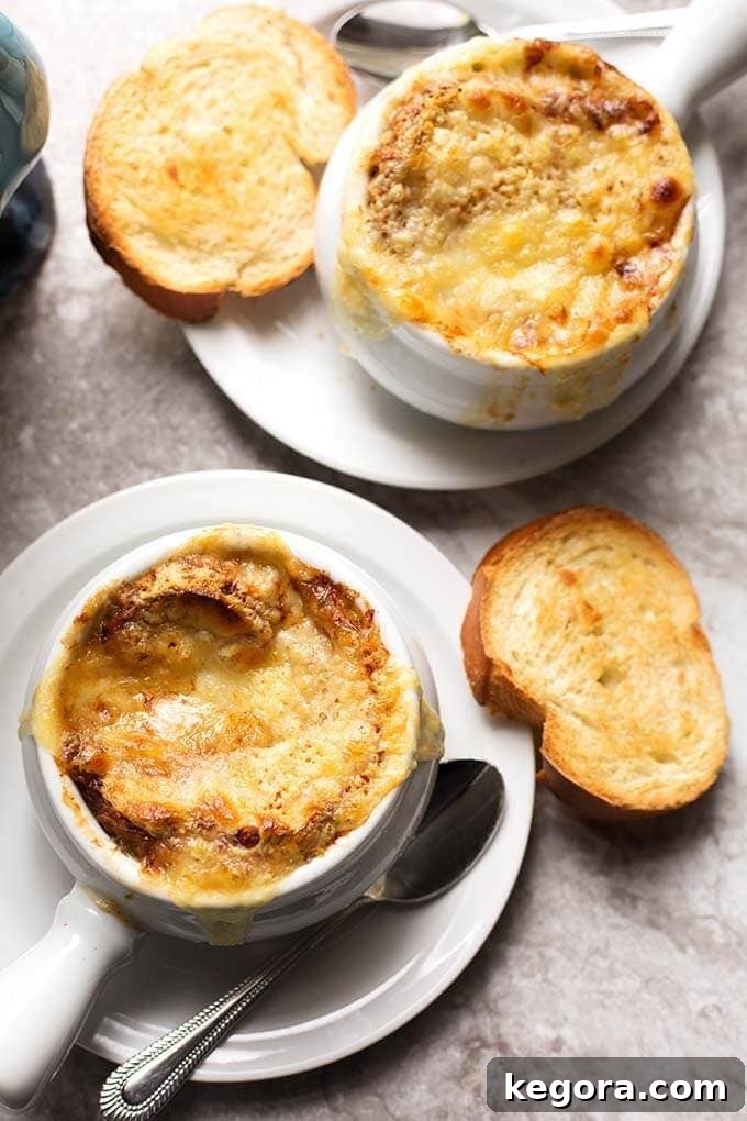 Top view of two bowls of French Onion Soup with melted Gruyere cheese and a small slice of toasted baguette on white plates, garnished with fresh herbs.