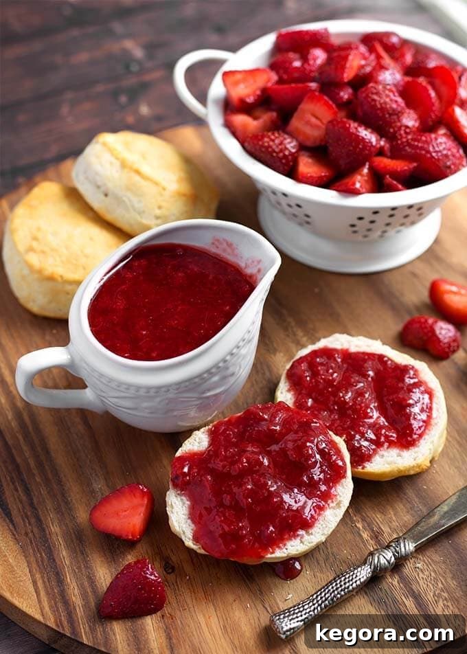 Strawberry Sauce in a small white pitcher next to biscuits with strawberry syrup spread on them next to a white colander full of halved strawberries