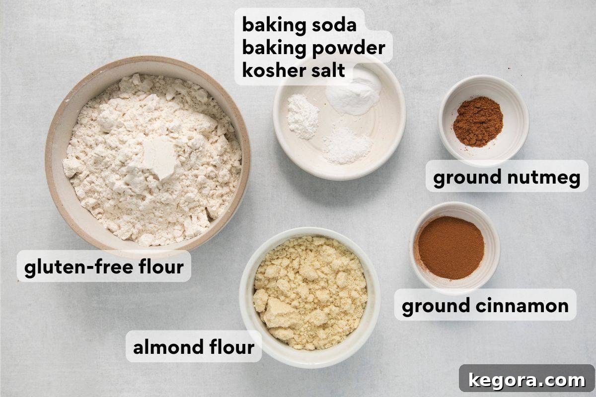 Various dry ingredients for applesauce bread, measured and laid out in separate bowls with labels for gluten-free flour, almond flour, baking soda, baking powder, salt, cinnamon, and nutmeg.