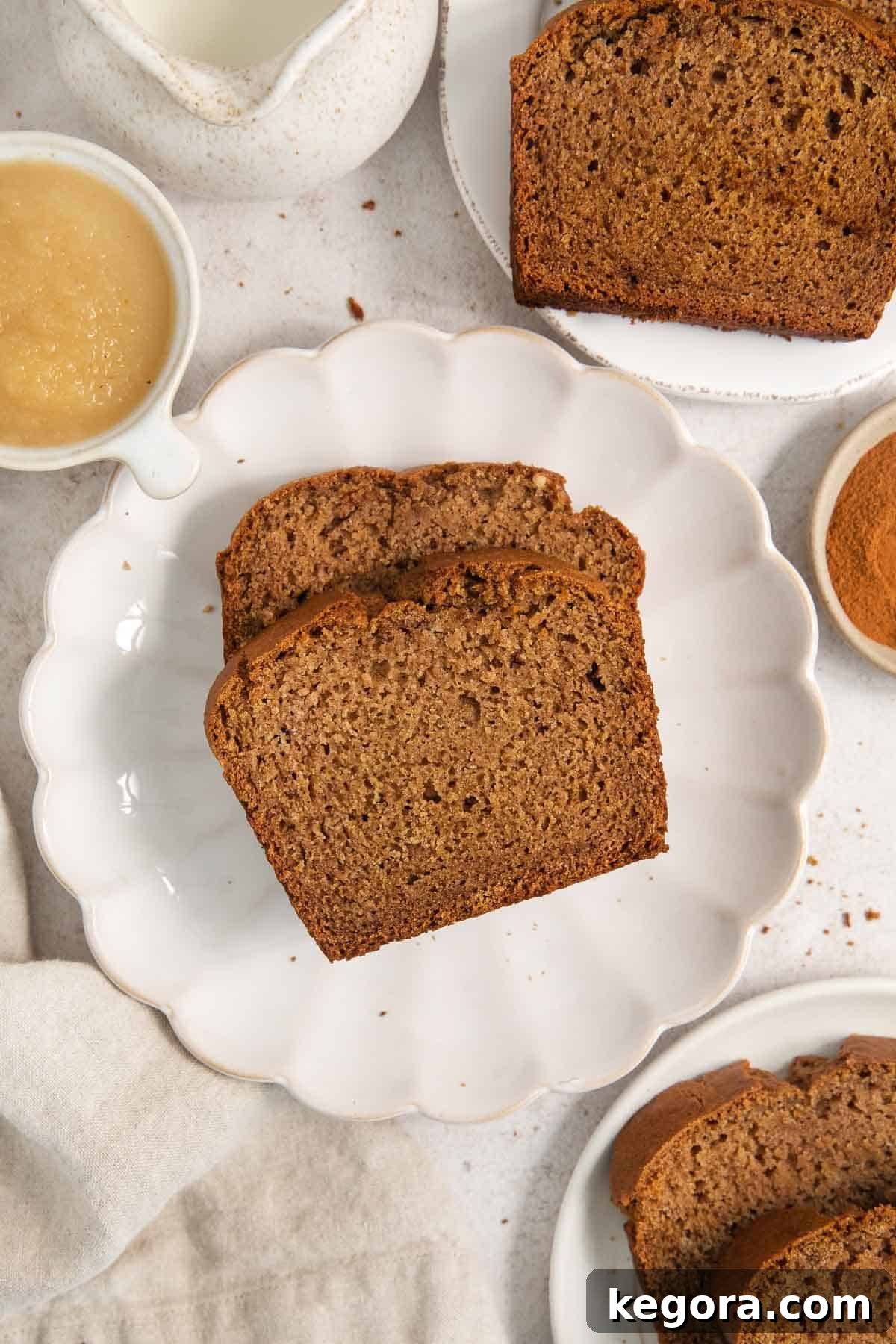 Two beautiful slices of gluten-free applesauce bread arranged on a white plate, surrounded by whole apples and a baked loaf, highlighting its enticing appearance.