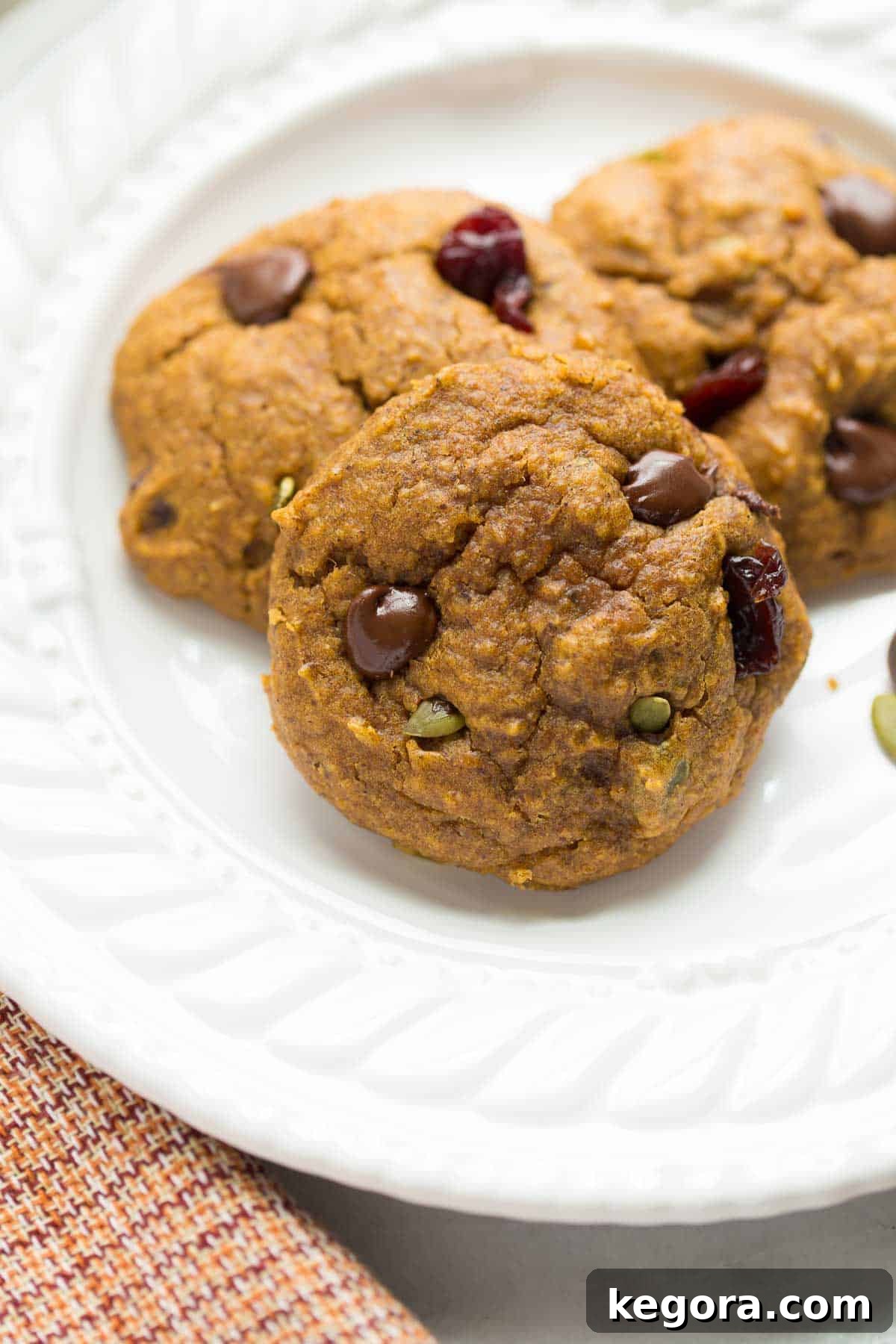 Cozy Vegan Pumpkin Breakfast Cookies 6 Three perfectly baked vegan pumpkin cookies artfully arranged on a white plate, accompanied by a cozy plaid red and orange napkin, ready to be enjoyed.
