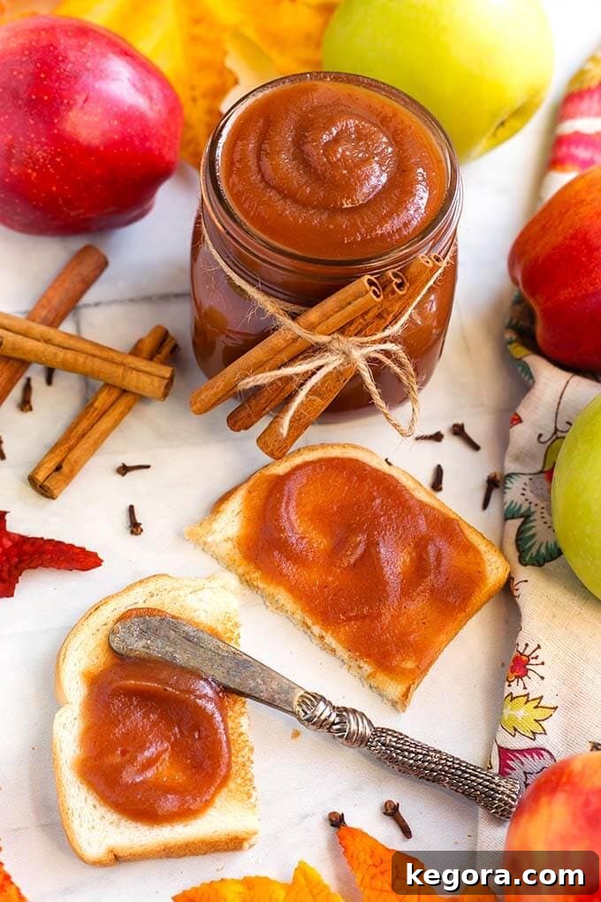 Quick Homemade Instant Pot Apple Butter 3 Toasted bread with Apple Butter spread on it with a silver knife in front of a Small glass jar of Apple Butter with cinnamon sticks tied with twine, all surrounded by cloves, cinnamon sticks, and apples