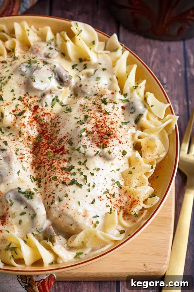 Close up of Creamy Chicken Breasts generously topping egg noodles in a beige bowl, placed on a rustic wooden board, highlighting the creamy sauce and tender chicken.