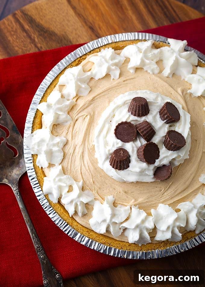 A beautifully presented No Bake Peanut Butter Pie, crowned with generous swirls of whipped cream and delightful chocolate candies. The pie rests in a convenient disposable pie tin, alongside a gleaming silver serving spoon, all elegantly arranged on a rustic wooden board, ready to be enjoyed.