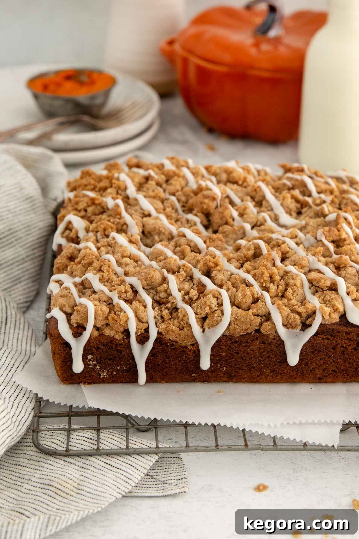 Gluten-Free Pumpkin Crumb Cake Recipe 2 A rectangular pumpkin crumb cake sits on a cooling rack, topped with streusel and white icing drizzle. A striped towel, plates, orange dish, and milk bottle are in the background.