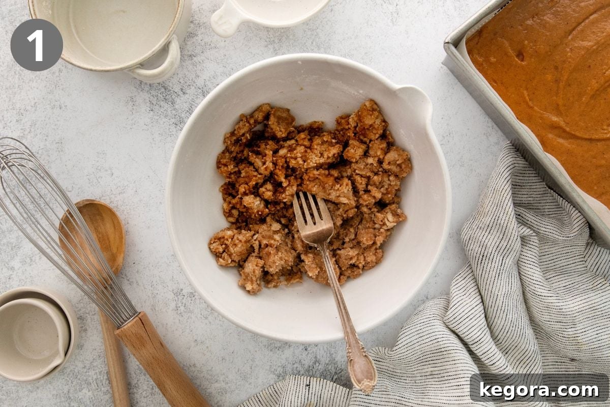 Gluten-Free Pumpkin Crumb Cake Recipe 4 A white bowl with crumb topping mixture and a fork sits on a counter, surrounded by a whisk, wooden spoon, empty cups, a striped cloth, and a pan of batter.