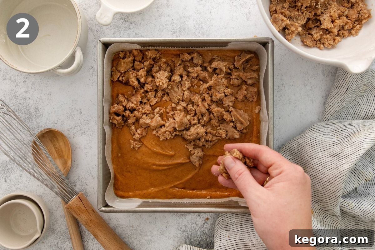 Gluten-Free Pumpkin Crumb Cake Recipe 5 A hand sprinkles crumb topping over pumpkin batter in a square pan. Nearby are mixing bowls, a whisk, a wooden spoon, and a striped towel on a light countertop.