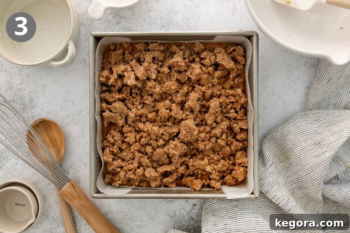 Gluten-Free Pumpkin Crumb Cake Recipe 6 A baking pan filled with a crumbly brown topping sits on a light surface surrounded by kitchen utensils, bowls, and a striped towel. A circle with the number 3 is in the upper left corner.