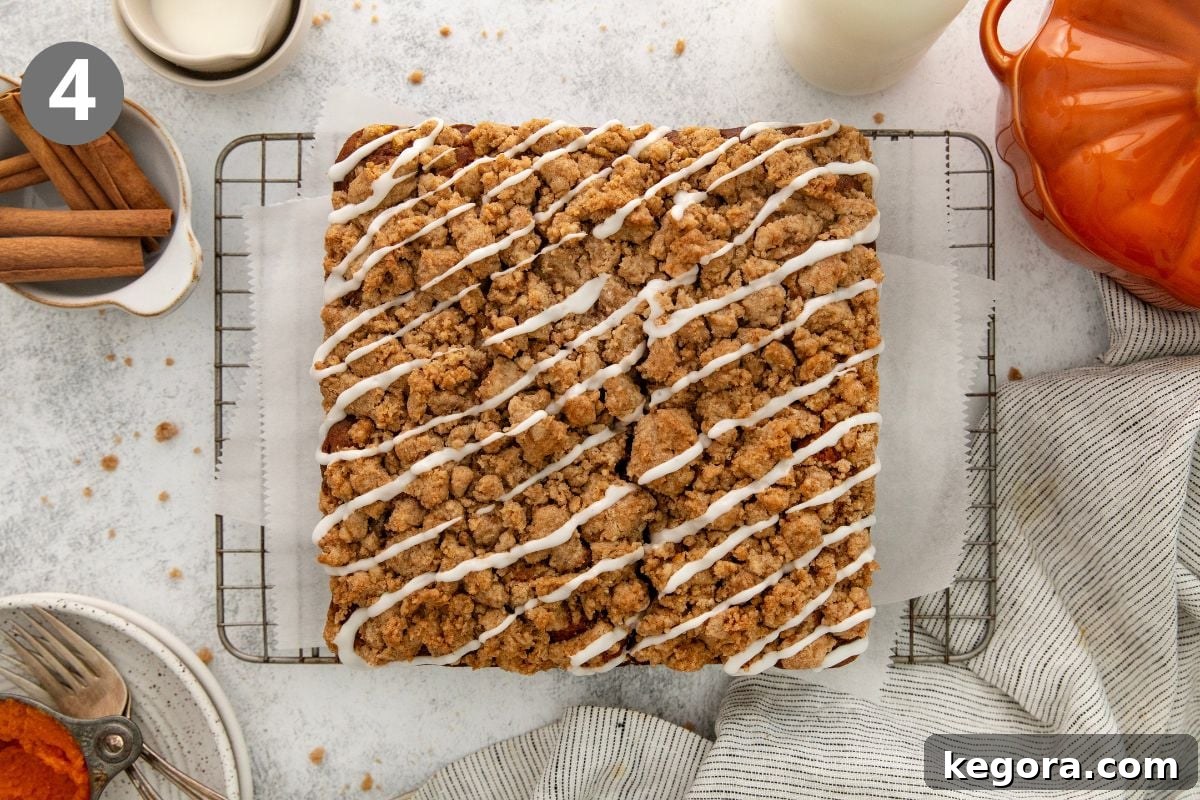 Gluten-Free Pumpkin Crumb Cake Recipe 7 A large rectangular crumb cake with white icing drizzle sits on a wire rack lined with parchment paper, surrounded by a glass of milk, cinnamon sticks, and a striped napkin.