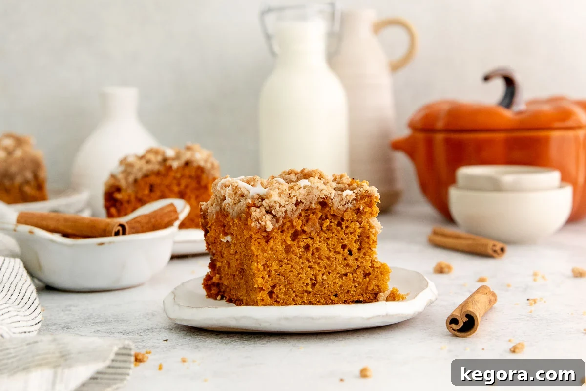 Gluten-Free Pumpkin Crumb Cake Recipe 8 A slice of pumpkin crumb cake with crumb topping on a plate, surrounded by cinnamon sticks, a milk bottle, and autumn-themed dishes in a cozy setting.