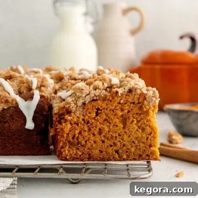 A close-up of a sliced pumpkin crumb cake with icing drizzle on a cooling rack. Milk and ceramic jugs are blurred in the background.