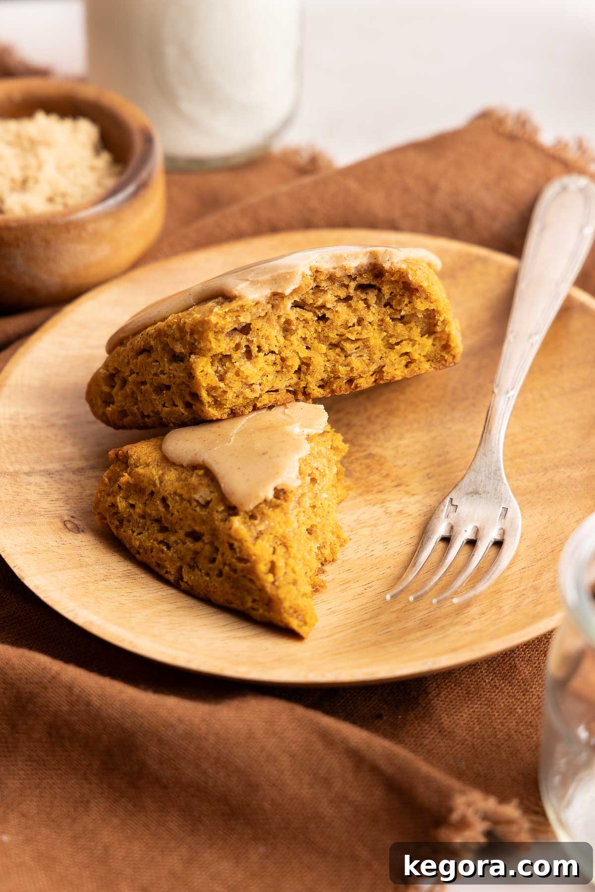 Flaky Gluten-Free Pumpkin Scones Recipe 2 A wooden plate holds two triangular pieces of pumpkin scone with brown sugar glaze, next to a fork. A bowl of brown sugar and a glass of milk are in the background on a brown cloth.