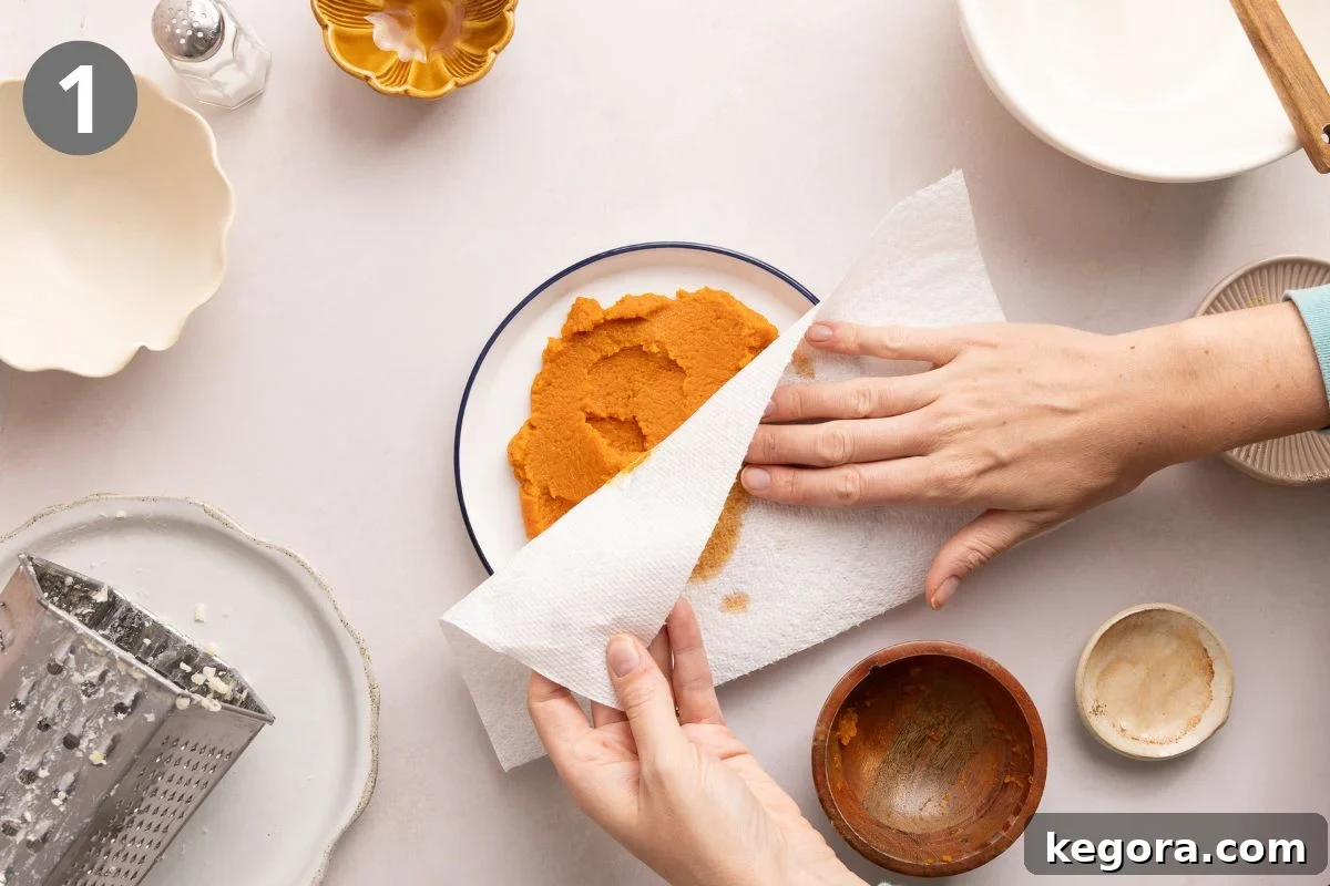 Flaky Gluten-Free Pumpkin Scones Recipe 4 A person presses a paper towel onto a plate of pumpkin puree on a white table, surrounded by various empty bowls and a grater.