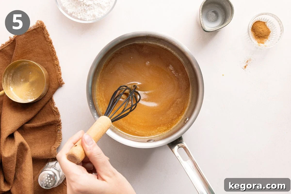 Flaky Gluten-Free Pumpkin Scones Recipe 8 A hand whisks a brown sugar glaze in a saucepan. Nearby are a brown cloth, a small gold bowl, flour, spices, and a salt shaker.