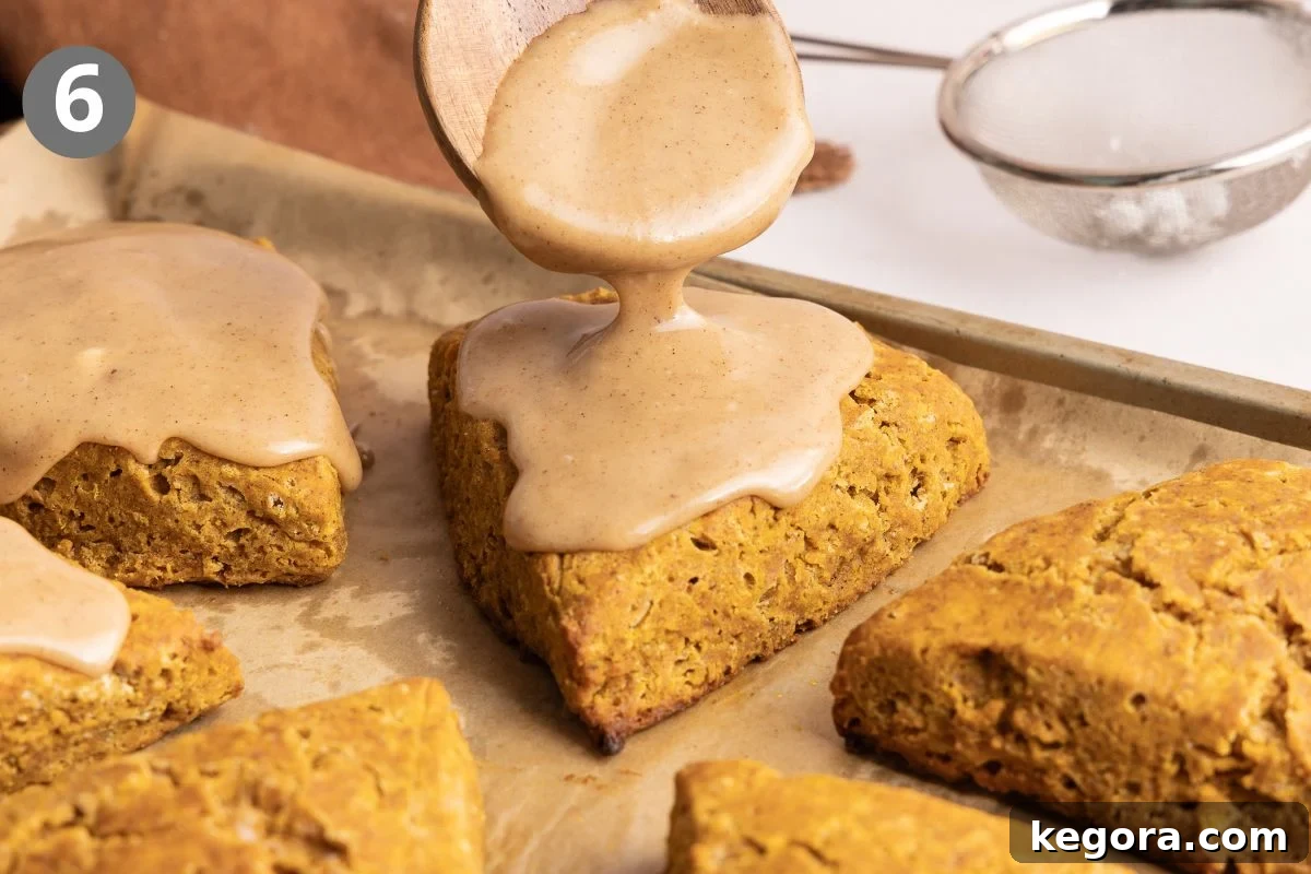 Flaky Gluten-Free Pumpkin Scones Recipe 9 A wooden spoon drizzles brown sugar glaze over a triangular scone on a baking sheet, surrounded by other scones.