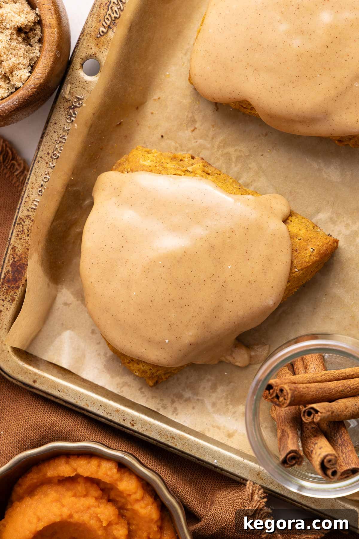 Flaky Gluten-Free Pumpkin Scones Recipe 10 Pumpkin scone topped with a brown sugar glaze on a baking sheet, with cinnamon sticks, brown sugar, and pumpkin puree nearby.