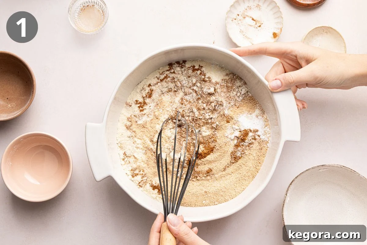 Hands holding a white mixing bowl with flour and brown sugar, using a whisk to combine the ingredients. Several empty bowls and dishes sit nearby on a light surface.