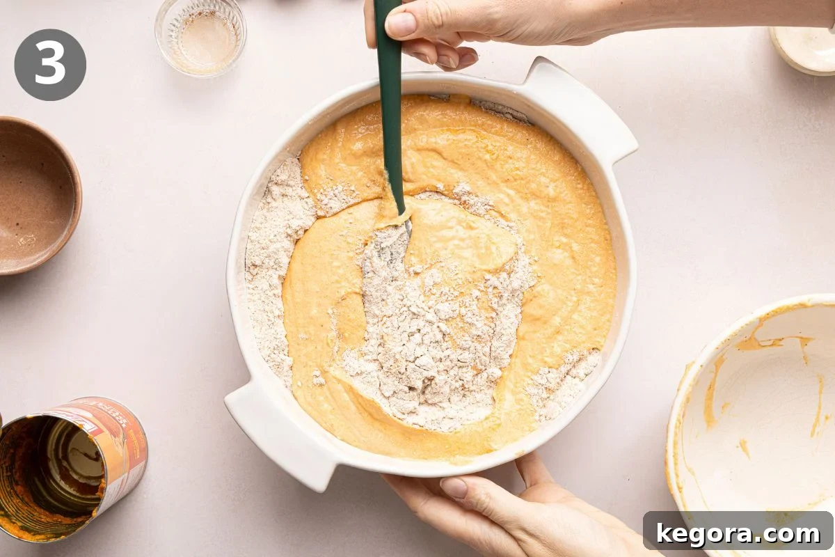 A person mixing flour into a yellow batter in a white bowl with a green spatula, step 3 in a recipe. Small bowls with ingredients and an open can are nearby on a light surface.