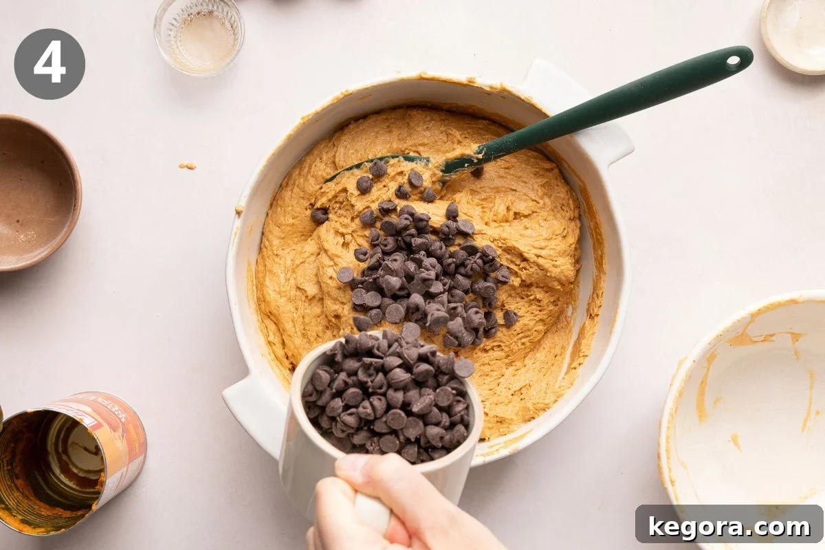 A hand pours chocolate chips from a white cup into pumpkin muffin batter in a mixing bowl with a green spatula. Several small bowls and an open can are nearby.