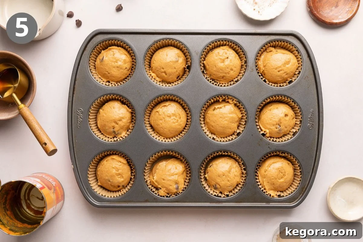 Pumpkin protein muffin batter in a non-stick muffin pan, surrounded by various baking utensils and empty bowls on a light countertop.