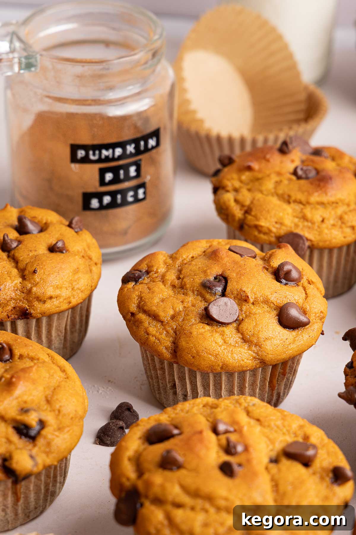 Pumpkin protein muffins on a white surface, with a glass jar labeled Pumpkin Pie Spice, an empty muffin liner, and a glass of milk in the background.
